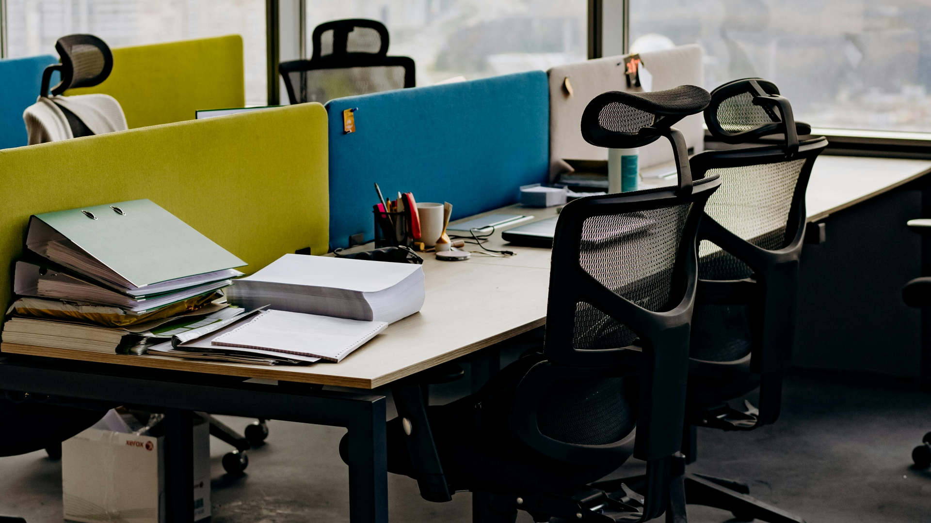 Postos de trabalho modernos num escritório, exemplificando a necessidade de análise ergonómica pela Jéssica Pereira Fisioterapia. A imagem mostra secretárias organizadas com cadeiras ergonómicas ajustáveis e divisórias coloridas, focando na configuração correta para prevenir lesões e promover o conforto laboral.