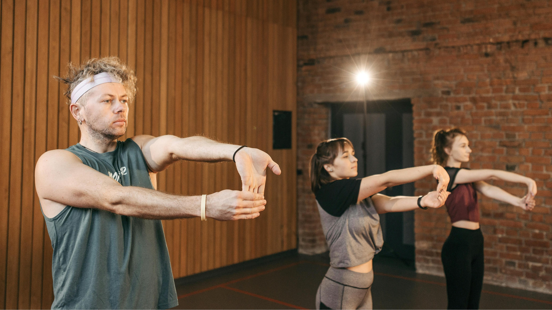 Grupo de colaboradores participa numa sessão de ginástica laboral orientada pela Jéssica Pereira Fisioterapia. A imagem mostra três pessoas a realizar exercícios de alongamento dos punhos e antebraços num ambiente de estúdio, visando a prevenção de lesões ocupacionais.